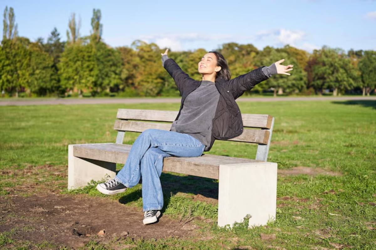  A woman sitting on a bench in a park, wearing jeans and footwear, with her arms outstretched. The scene includes grass, trees, and a clear sky in the background.