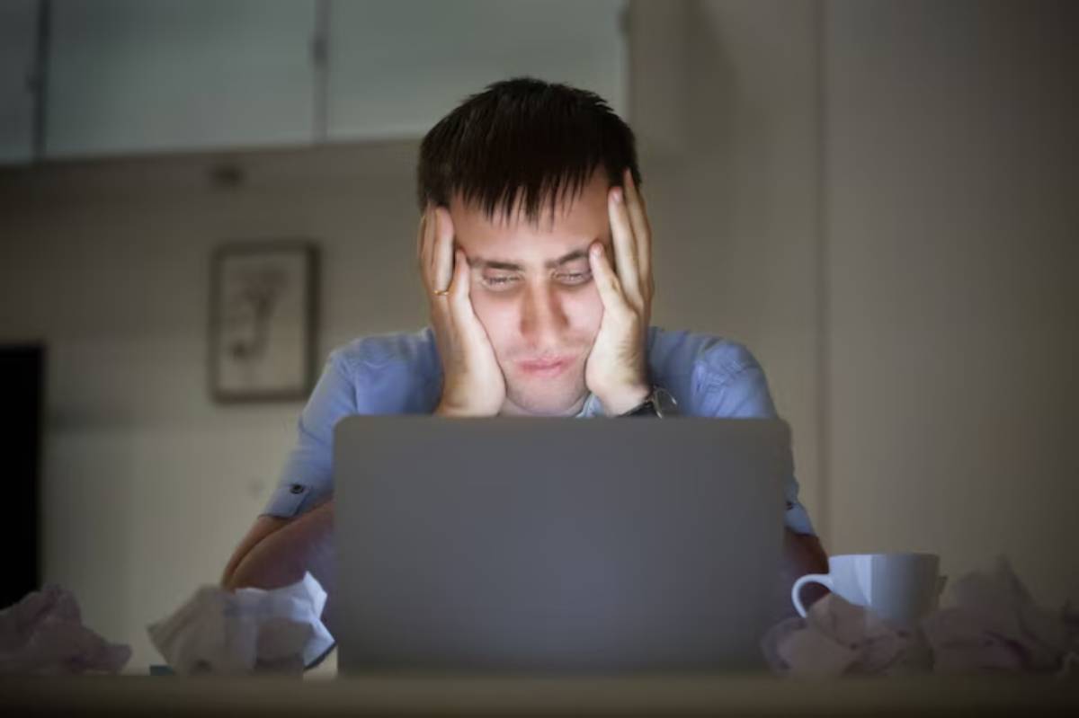  A young man sitting indoors, looking at a laptop with his hands on his face, appears to be deep in thought.