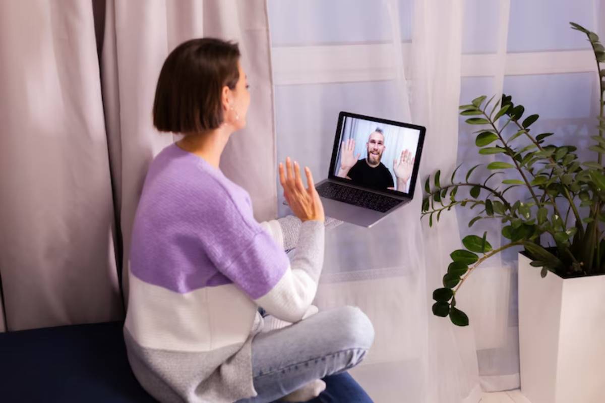 A woman sits on a couch, smiling and waving at a person on a laptop screen, with a plant nearby and soft curtains in the background. 