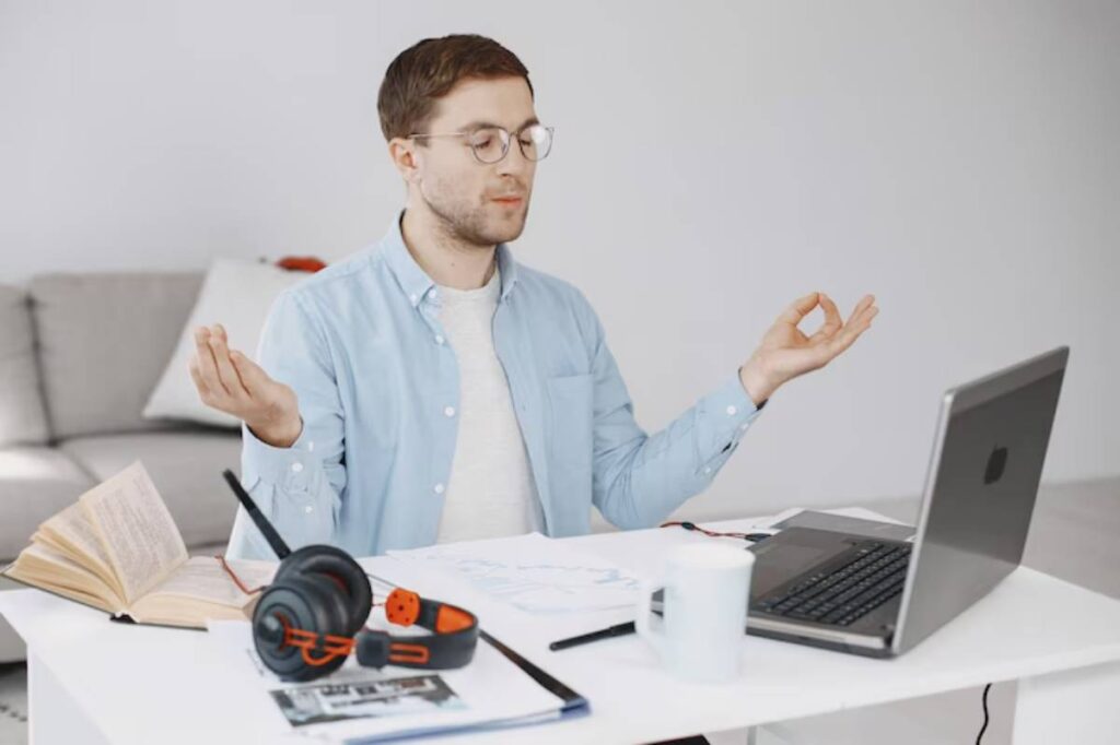 A person in a light blue shirt meditates in front of a laptop, with headphones, a mug, and an open book on the table.