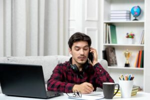 A person in a plaid shirt sits at a desk with a laptop, holding a phone, surrounded by stationery and books in a cozy room.