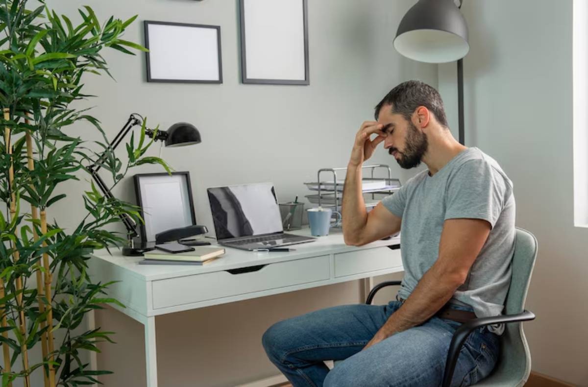  A man sits in a home office, with a laptop and desk lamp, looking stressed while resting his forehead on his hand.