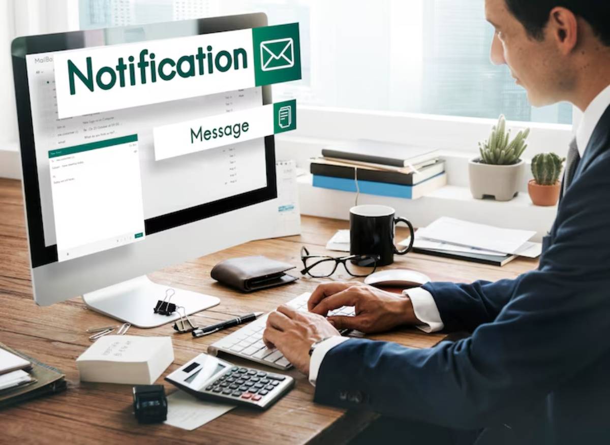 A person in a suit works at a desk with a computer displaying email notifications, surrounded by office supplies and plants. 