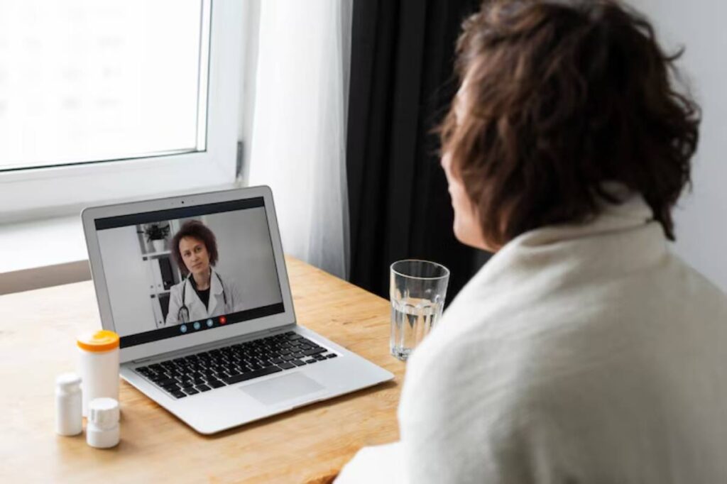 A person in a white robe is engaged in a video call with a doctor on a laptop, accompanied by a glass of water and medication bottles.