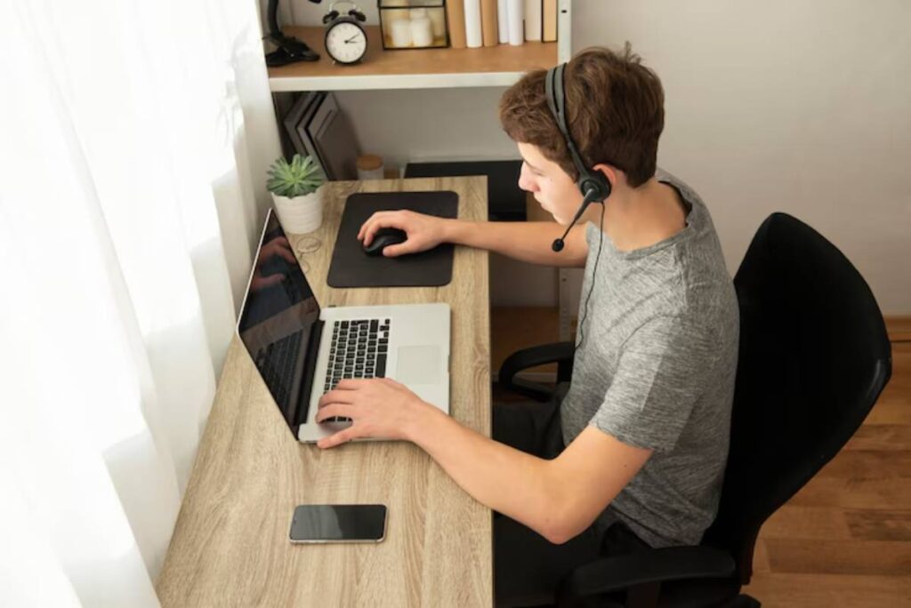 A person wearing a headset sits at a desk with a laptop and smartphone, focused on their work in a well-lit workspace.