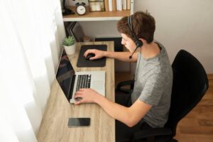 A person wearing a headset sits at a desk with a laptop and smartphone, focused on their work in a well-lit workspace.