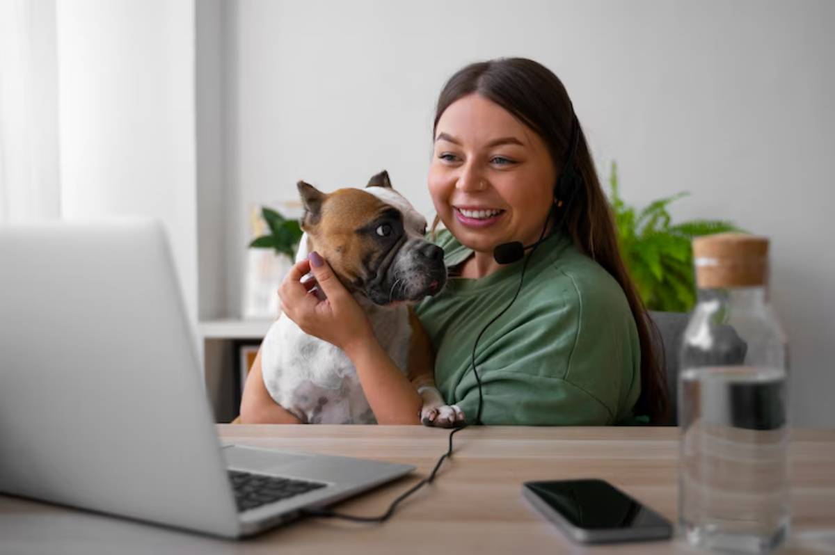 A relaxed person wearing a green shirt holds a dog, seated at a desk with a laptop, a water bottle, and a smartphone nearby. 
