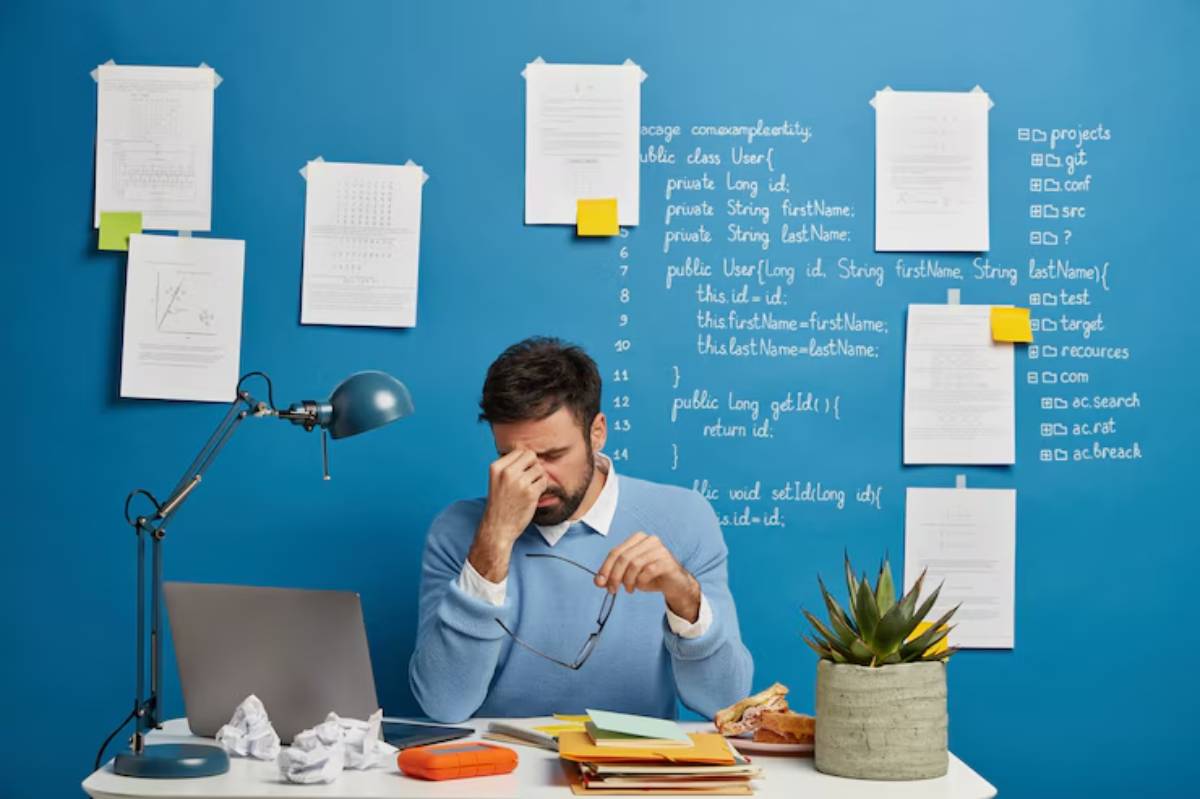  A person sits at a desk with papers and a laptop, surrounded by notes and code on a blue wall, with a plant and snacks nearby.