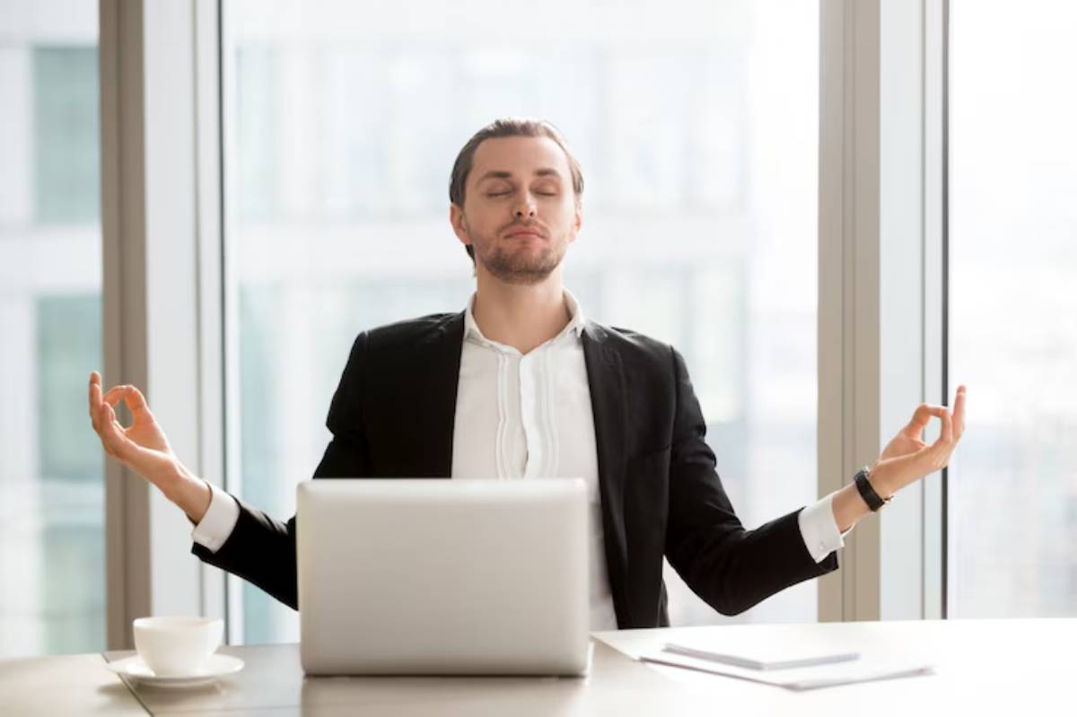  A person in business attire sits at a desk with a laptop, performing a meditative gesture in a bright, modern office setting.