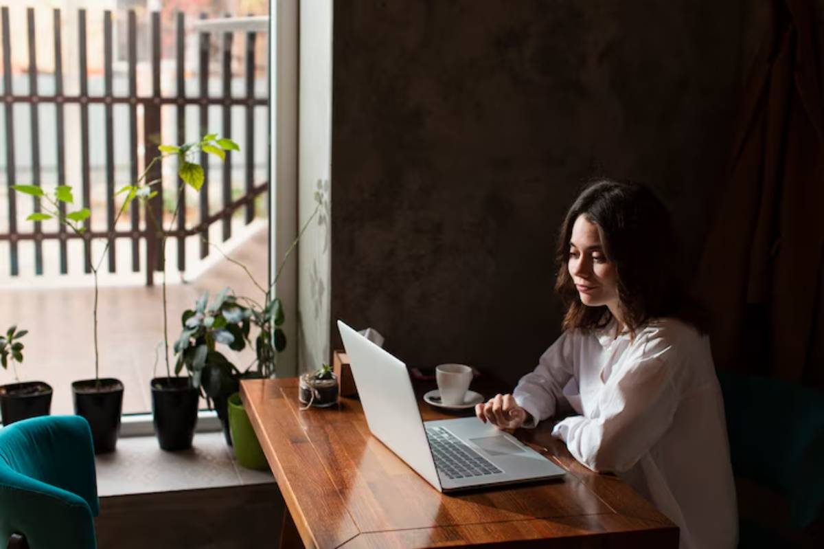  A person sits at a wooden table with a laptop, surrounded by potted plants and a window revealing a fenced outdoor view.