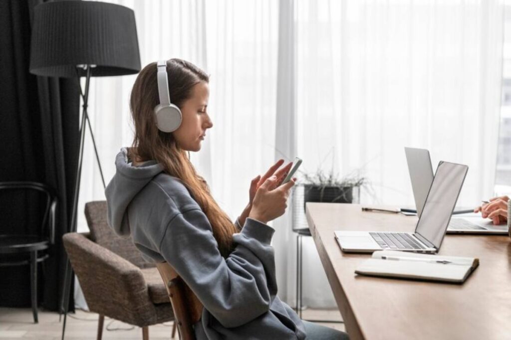 A person wearing headphones sits at a table, using a smartphone, with laptops and a plant visible in a bright, cozy workspace.