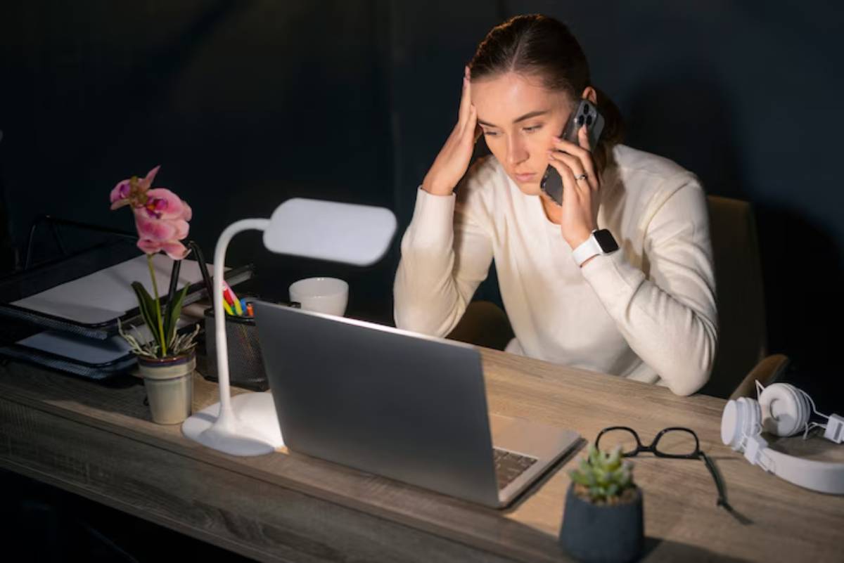  A person in a white sweater holds a phone, looking stressed at a wooden desk with a laptop, plants, and office supplies.