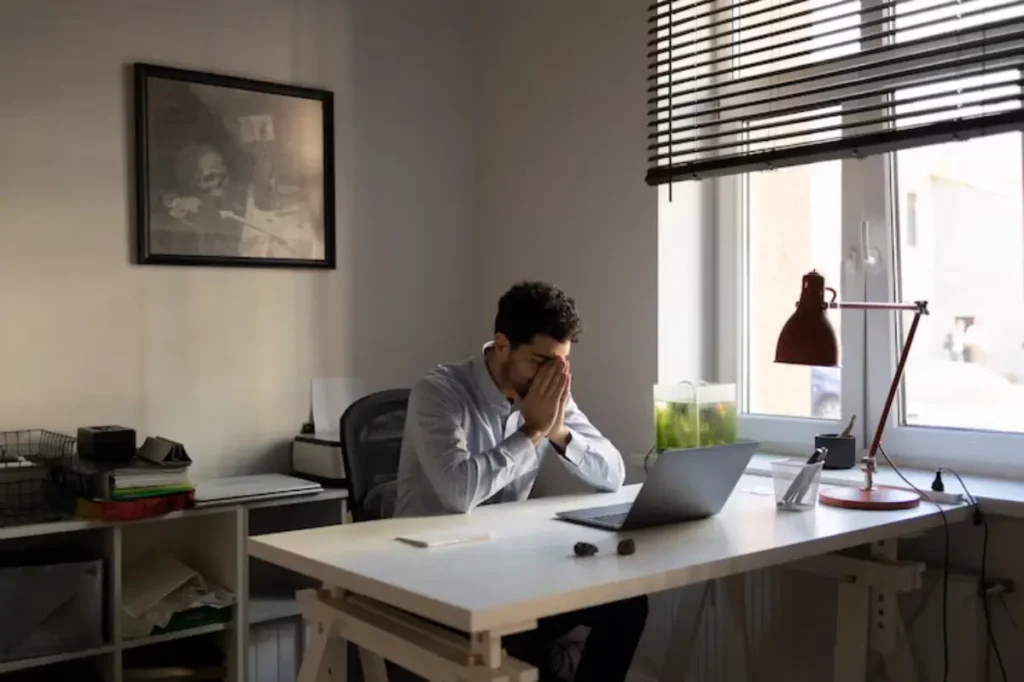 A person sits at a desk in a bright room, with a laptop, a green fish tank, and a desk lamp, deep in thought. A framed picture adorns the wall.