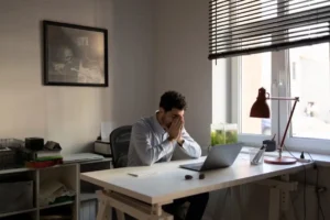 A person sits at a desk in a bright room, with a laptop, a green fish tank, and a desk lamp, deep in thought. A framed picture adorns the wall.