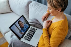 A person sits on a couch, using a laptop for a video call, wearing a cozy mustard sweater. Soft furnishings and natural light create a warm ambiance.