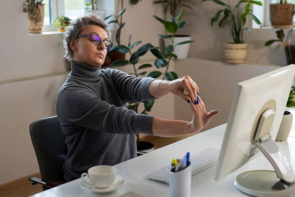 A person in a gray sweater stretches their hands at a desk with a computer, surrounded by indoor plants and a coffee cup.