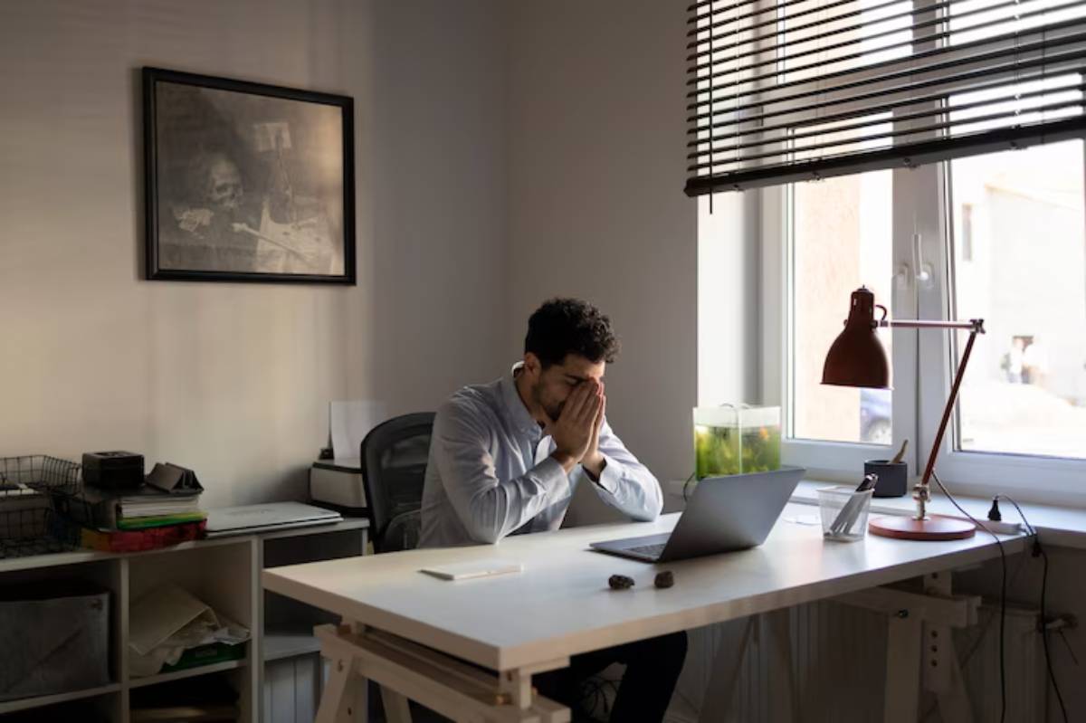  A person sits at a desk, hands clasped in front of their face, in a well-lit, modern office with a laptop and a fish tank nearby.