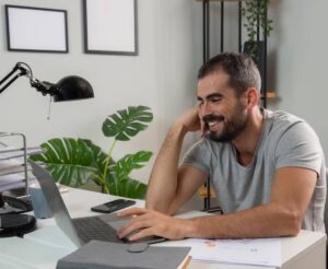 A person in a gray t-shirt sits at a desk with a laptop, focusing on work, surrounded by plants and stationery items.