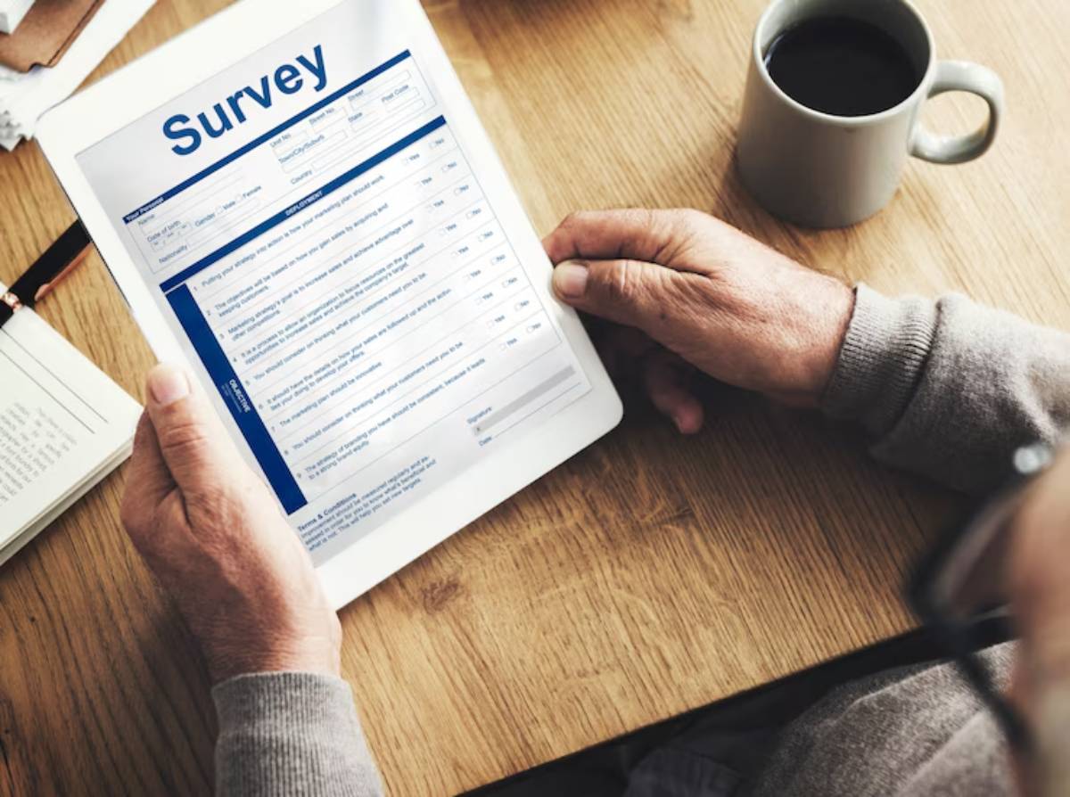  An elderly person holds a tablet displaying a survey form, with a coffee cup and notebook on a wooden table.