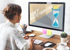 A person working at a desk, viewing a computer screen with the message, Don't waste your time, alongside an hourglass and dollar sign.