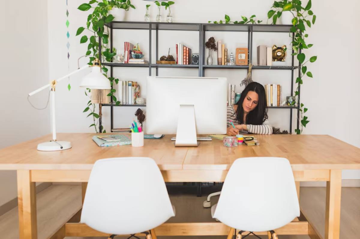  A stylish, modern workspace featuring a wooden table, a computer monitor, stationery, and a vibrant bookshelf adorned with plants.