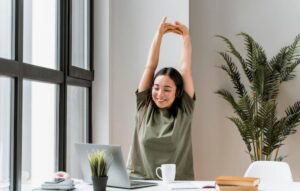 A person wearing headphones stretches their arms above their head while seated at a desk with a laptop and indoor plants.