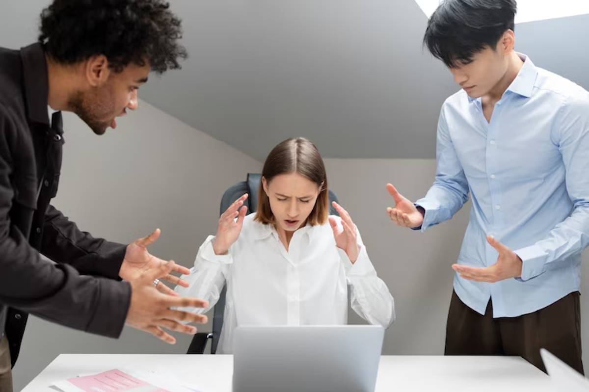  A woman seated at a desk appears stressed while two individuals engage animatedly, gesturing as they discuss something serious.