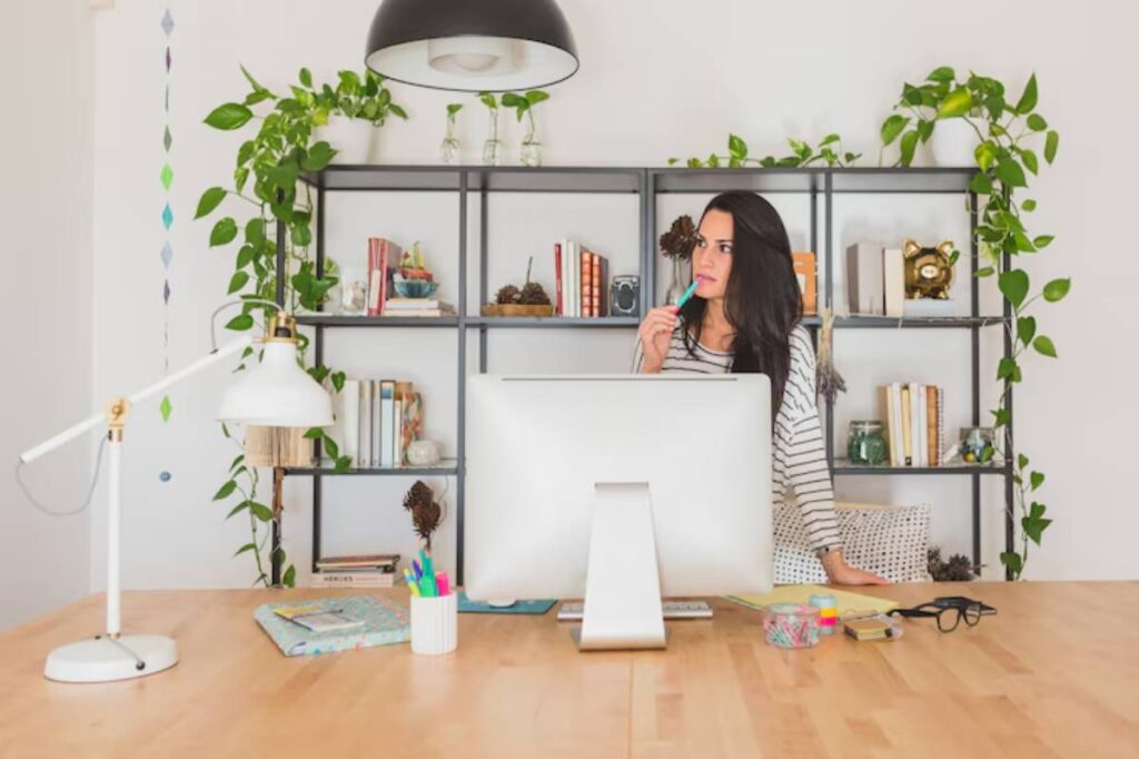 A woman stands at a desk with a computer, surrounded by plants and bookshelves filled with various items, including stationery and decor.