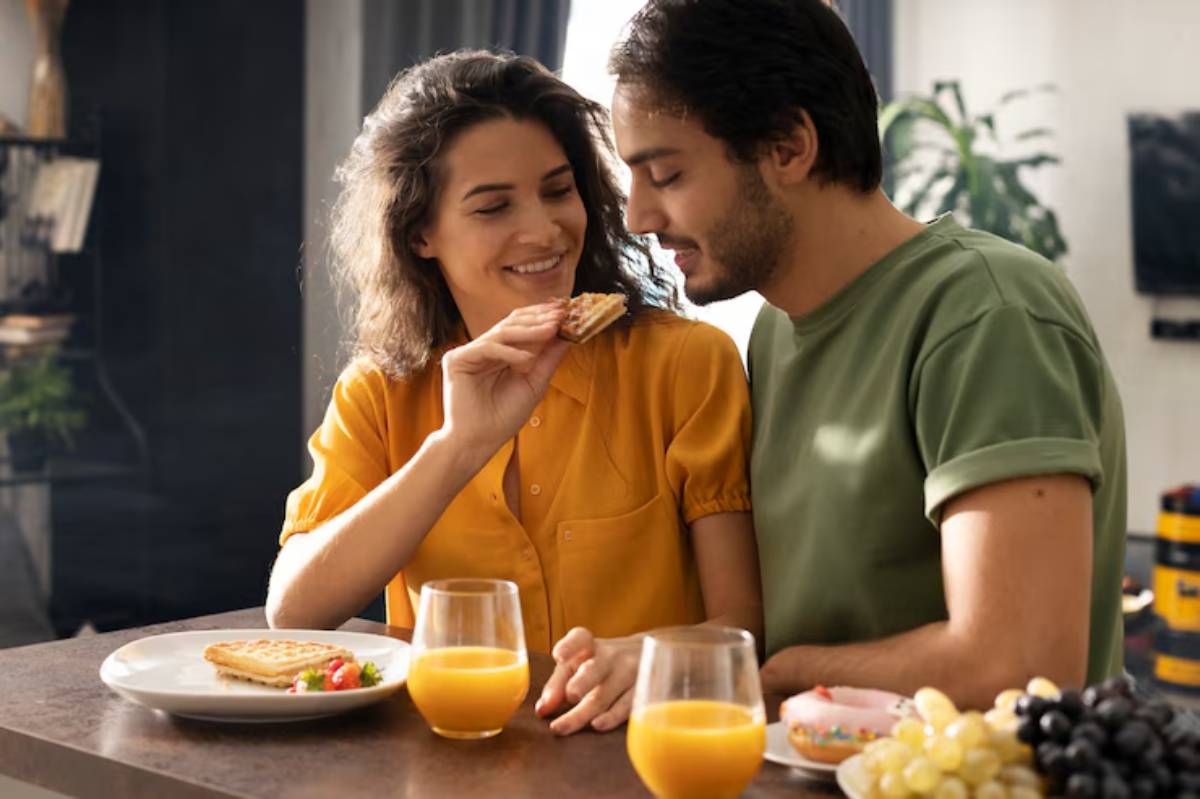 A man and woman sitting at a table indoors, enjoying breakfast together. The table is set with food and drinks, including orange juice. They are casually dressed and smiling as they eat. 